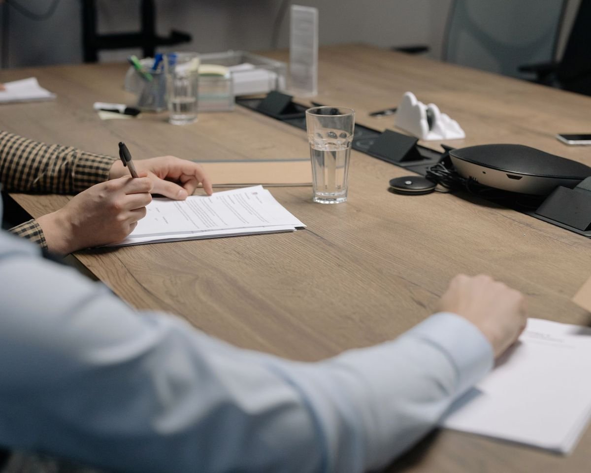 Duas pessoas assinando um documento sobre a mesa de uma sala de reunião
