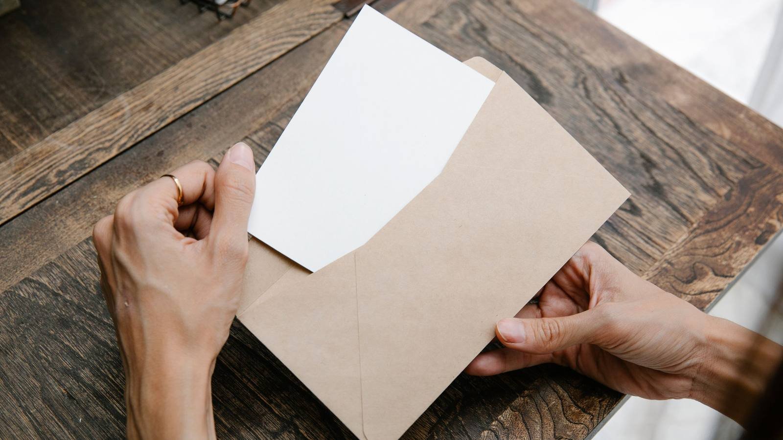 Mãos retirando uma folha de dentro de um envelope pardo, sobre mesa de madeira