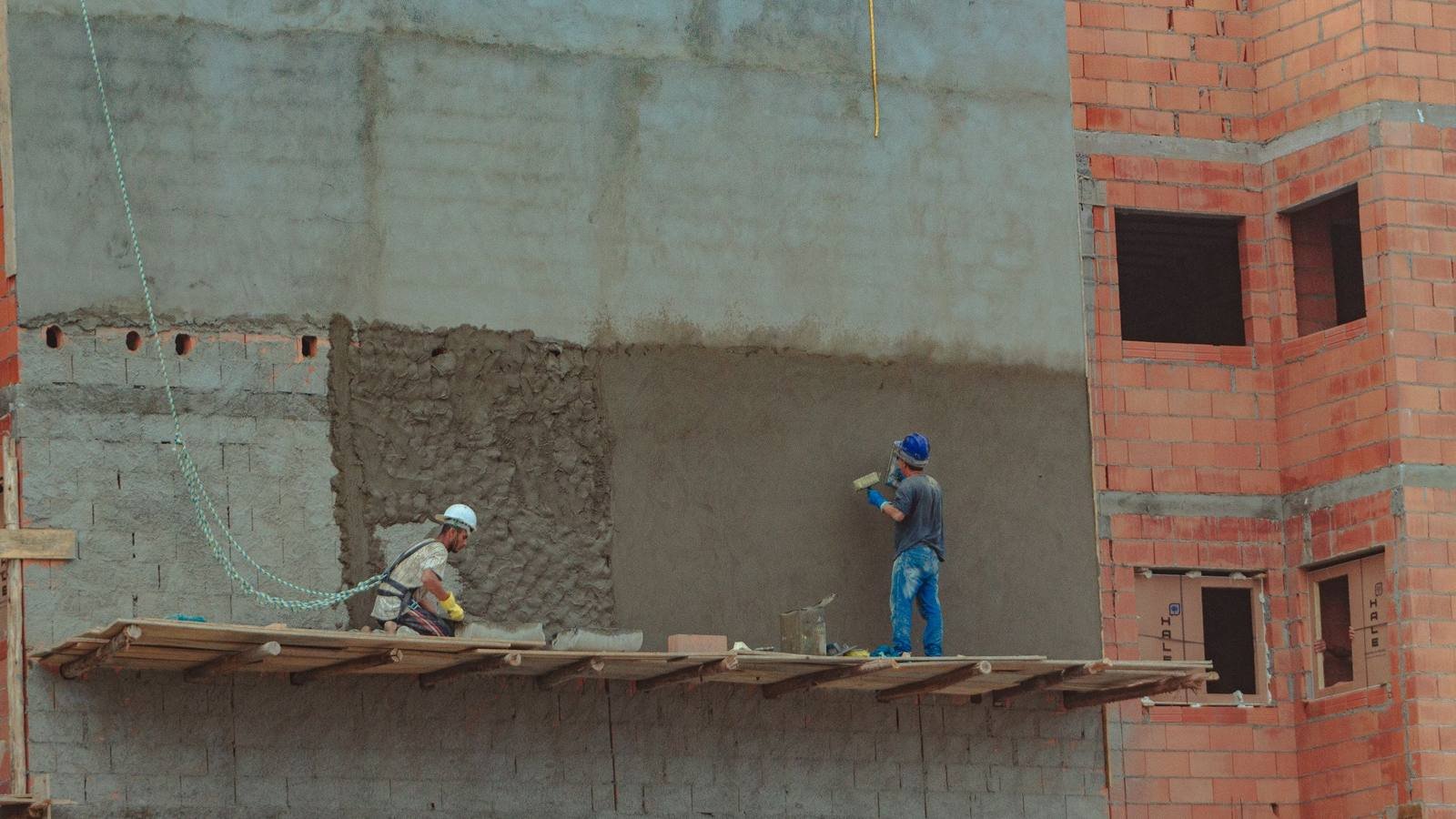 Canteiro de obra com dois operários trabalhando em uma parede de tijolo cerâmico